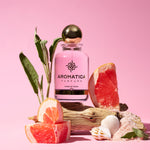 Aromatica Parfums Eau de Parfum bottle with a pink liquid and a metallic gold cap, styled with grapefruit slices, green leaves, driftwood, and seashells on a solid pink background.