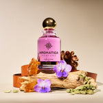 A bottle of Aromatica Perfums Eau de Parfum with a pink label and a gold spherical cap, styled on a rustic log with purple flowers, green cardamom pods, amber crystals, and a pine cone against a warm beige background.