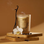 Aromatica vanilla bean candle with vanilla pods and white orchids on a wooden stand against a brown background.